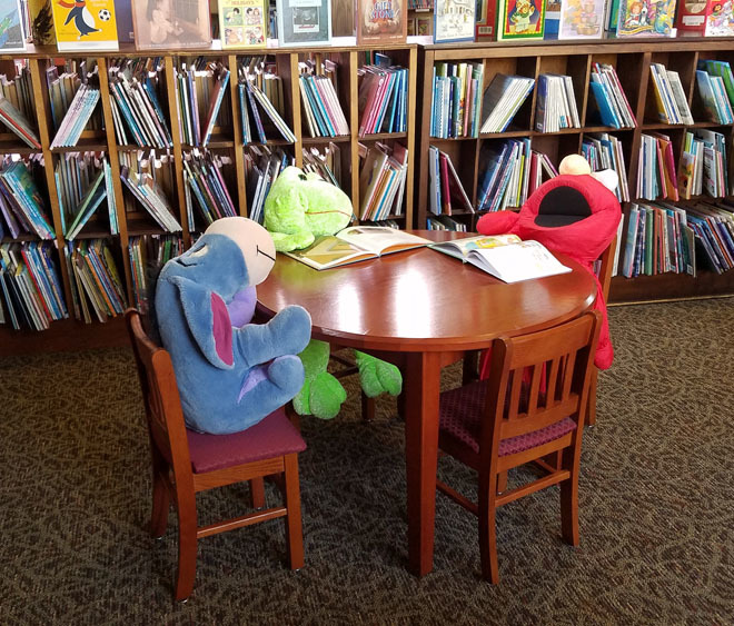 a small round table with four chairs. in three chairs is a stuffed animal of eeyore, elmo, and a frog.